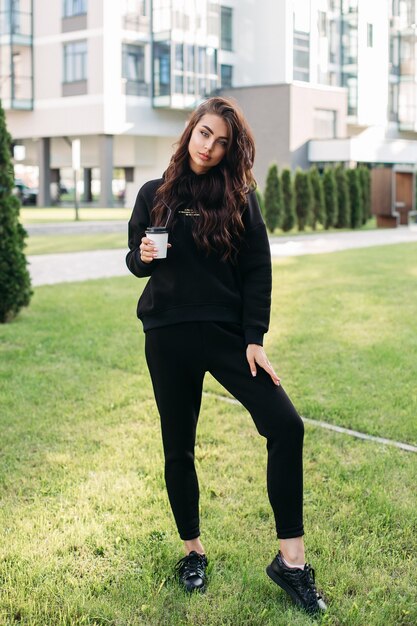 Bellissimo modello femminile con capelli ricci che indossa una tuta sportiva nera elegante e tiene in mano una tazza di caffè. Moda femminile. Stile di vita della città