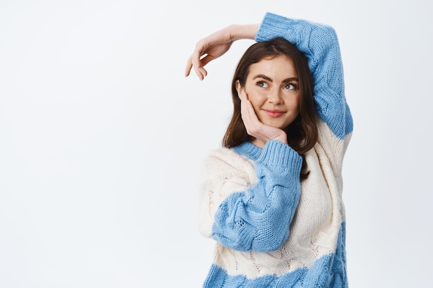 Bellissimo modello femminile in maglione caldo, toccando il viso naturale con un trucco leggero e tenendo la mano rilassata sulla testa su bianco