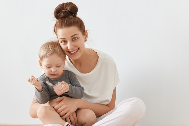 Bellissimo ritratto di giovane madre e bambino seduti insieme sul muro bianco. Felice femmina caucasica con un mazzo di capelli castani in abiti bianchi tenendo il bambino in braccio, sorridendo sinceramente.