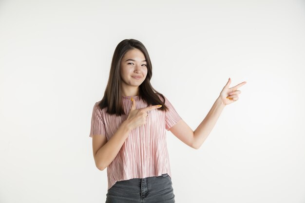 Bellissimo ritratto femminile a mezzo busto isolato su sfondo bianco studio. Giovane donna emotiva in abiti casual. Emozioni umane, concetto di espressione facciale. Indicando di lato, sorridendo.