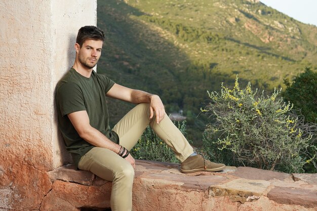 Bellissimo scatto di un affascinante maschio seduto e appoggiato al muro con una vista della natura dietro