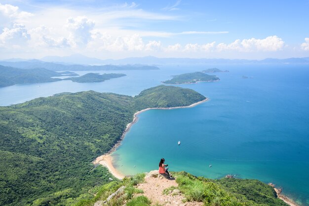 Bellissimo scatto di una donna seduta su una scogliera con un paesaggio di colline boscose e un oceano blu