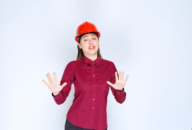 Bello architetto femminile in casco duro rosso che esamina macchina fotografica sopra la parete bianca.