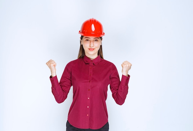 Bello architetto femminile in casco duro rosso che esamina macchina fotografica sopra la parete bianca.