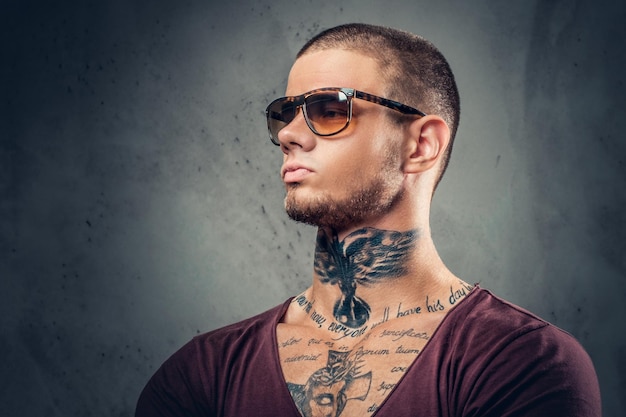 Bello, atletico maschio in occhiali da sole con tatuaggi sulle braccia e sul collo in posa su sfondo artistico grigio in uno studio.