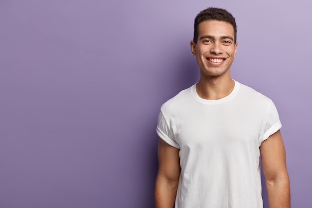 Bello e allegro giovane sportivo ha un corpo sportivo, braccia muscolose, indossa una maglietta bianca finta, ha i capelli corti e scuri, ha un sorriso accattivante a trentadue denti, si erge su un muro viola, copia spazio vuoto da parte