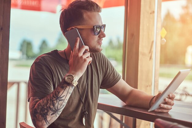 Bello ed elegante hipster si siede a un tavolo in un caffè lungo la strada, parlando al telefono e tiene il tablet.