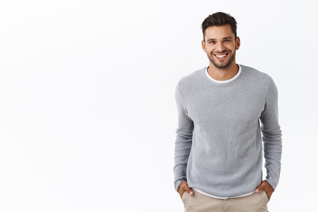 Bello ed elegante ragazzo ispanico moderno con barba alla moda taglio di capelli indossare maglione grigio sopra la maglietta tenere le mani in tasca e sorridere gioiosamente ridendo chiacchierando con gli amici sfondo bianco