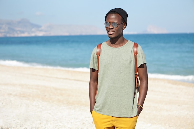 Bello felice giovane turista maschio nero con zaino vestito in abiti eleganti in piedi su una spiaggia di ciottoli con mare blu e cielo all'orizzonte, in attesa di amici per fare una bella passeggiata lungo la riva