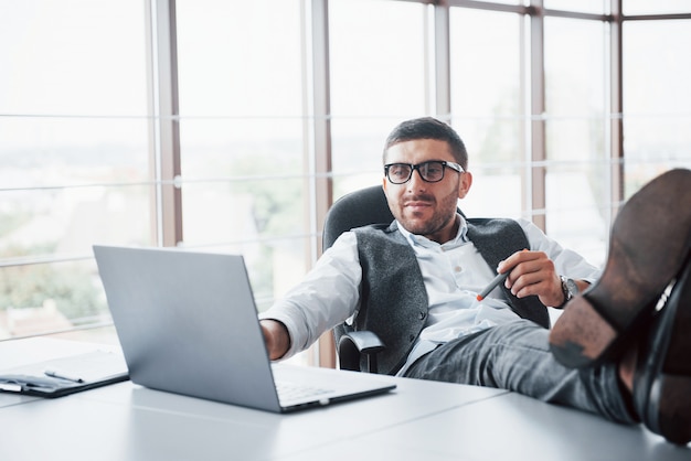 Bello giovane uomo d'affari con i vetri che tengono le sue gambe sul tavolo che esamina un computer portatile nell'ufficio