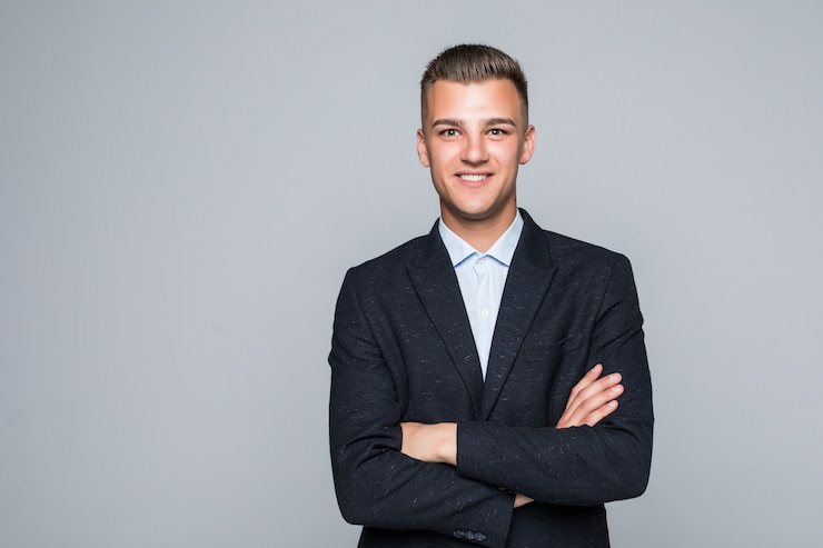 Foto gratuito bello giovane uomo d'affari studente in giacca tiene le braccia incrociate isolato sul muro grigio chiaro