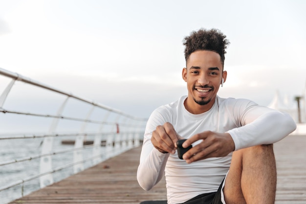 Bello giovane uomo riccio dalla pelle scura in maglietta a maniche lunghe sorride dolcemente e si siede su un tappetino da yoga vicino al mare Cool guy tiene le cuffie wireless