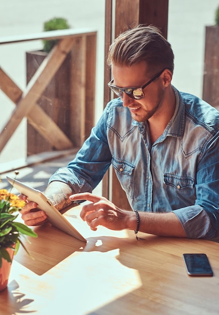 Bello hipster con un taglio di capelli alla moda e la barba si siede a un tavolo in un caffè lungo la strada, sembra qualcosa nel tablet.