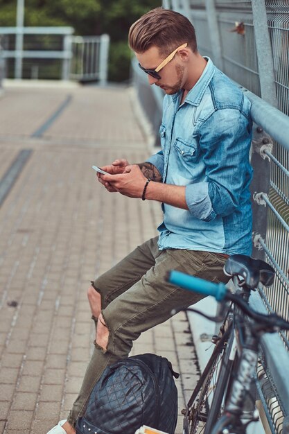Bello hipster con un taglio di capelli alla moda in occhiali da sole che riposa dopo essere andato in bicicletta, utilizzando uno smartphone.