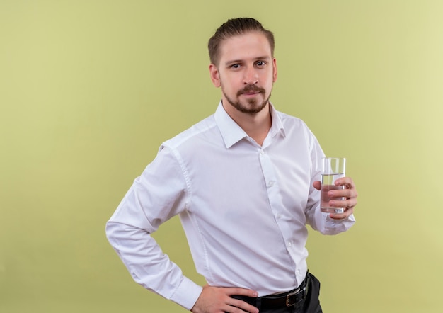 Bello imprenditore in camicia bianca bicchiere d'acqua che guarda l'obbiettivo con espressione fiduciosa in piedi su sfondo verde oliva