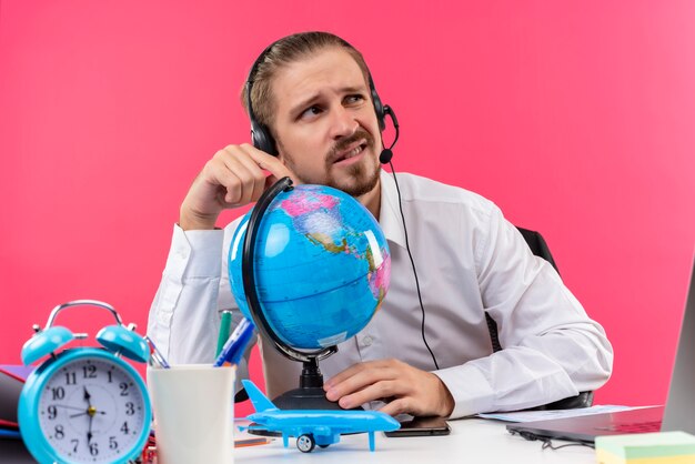 Bello imprenditore in camicia bianca con cuffie toccando il globo guardando da parte confuso seduto al tavolo in ufficio su sfondo rosa