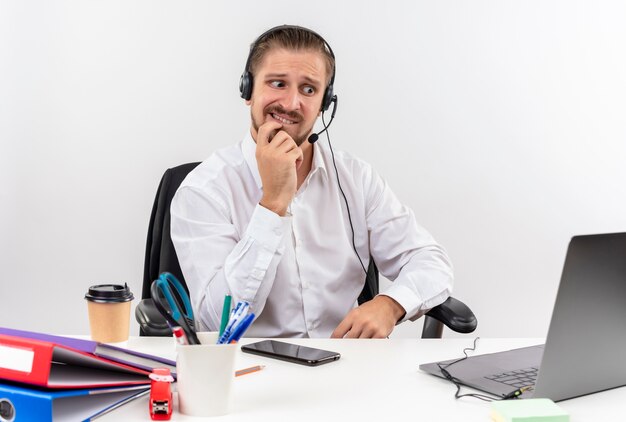 Bello imprenditore in camicia bianca e cuffie con un microfono che lavora su un laptop stressato e nervoso seduto al tavolo in offise su sfondo bianco