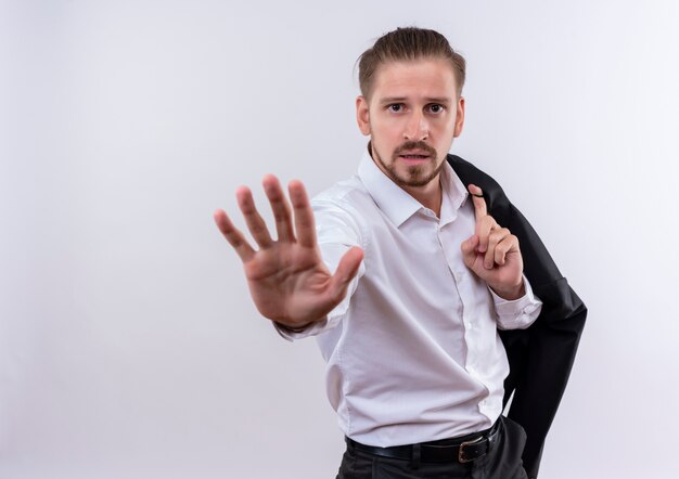 Bello imprenditore portando la sua giacca sulla spalla facendo il segnale di stop con la mano guardando con la faccia seria in piedi su sfondo bianco