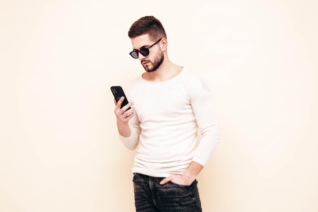 Bello modello fiducioso Uomo sexy ed elegante vestito con maglione e jeans Moda hipster maschio in posa vicino al muro bianco in studio Holding smartphone Guardando lo schermo del cellulare Usando app isolate