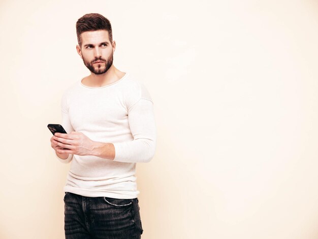 Bello modello fiducioso Uomo sexy ed elegante vestito con maglione e jeans Moda hipster maschio in posa vicino al muro bianco in studio Holding smartphone Guardando lo schermo del cellulare Usando app isolate