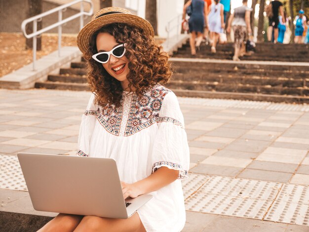 Bello modello sorridente con l'acconciatura dei riccioli di afro in vestito e cappello bianchi dai pantaloni a vita bassa di estate.