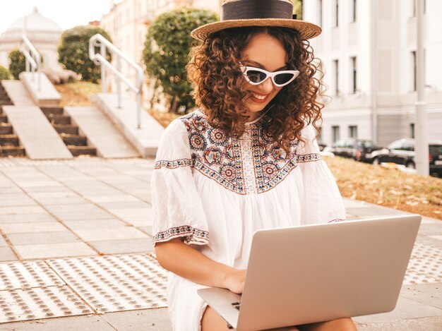 Bello modello sorridente con l'acconciatura dei riccioli di afro in vestito e cappello bianchi dai pantaloni a vita bassa di estate.