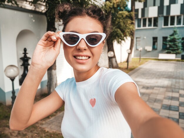 Bello modello sorridente con l'acconciatura di corna vestita in abbigliamento casual estivo Ragazza sexy spensierata in posa in strada in occhiali da sole. Scattare foto di autoritratto di selfie su smartphone