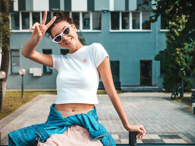 Bello modello sorridente con l'acconciatura di corna vestita in abiti jeans giacca estiva pantaloni a vita bassa Ragazza spensierata sexy in posa in strada. Donna divertente e positiva di tendenza divertendosi in occhiali da sole