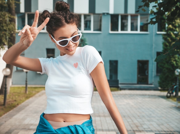 Bello modello sorridente con l'acconciatura di corna vestita in abiti jeans giacca estiva pantaloni a vita bassa Ragazza spensierata sexy in posa in strada. Donna divertente e positiva di tendenza divertendosi in occhiali da sole