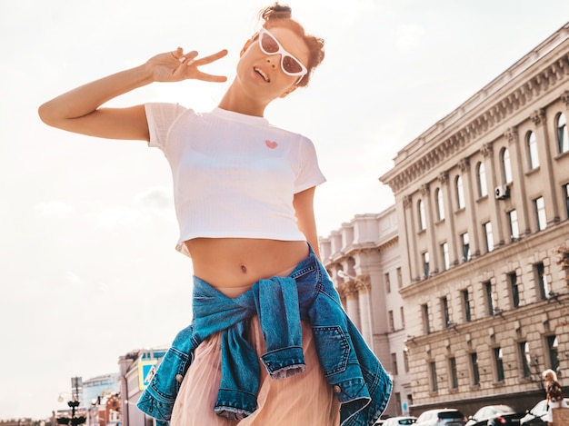 Bello modello sorridente con l'acconciatura di corna vestita in abiti jeans giacca estiva pantaloni a vita bassa Ragazza spensierata sexy in posa in strada La donna divertente e positiva alla moda mostra il segno di pace