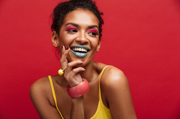 Bello ritratto del modello femminile afroamericano felice in camicia gialla che sorride e che posa sulla macchina fotografica, isolato sopra la parete rossa