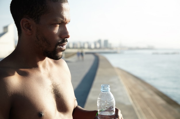 Bello scatto dell'afroamericano seminudo che è stanco del caldo estivo nelle grandi città. Giovane uomo rimandare i vestiti e in possesso di una bottiglia d'acqua