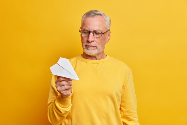 Bello serio saggio uomo maturo dai capelli grigi detiene aeroplani di carta fatti a mano andando a implementare l'idea vestita in maglione casual e occhiali isolati sopra il muro giallo