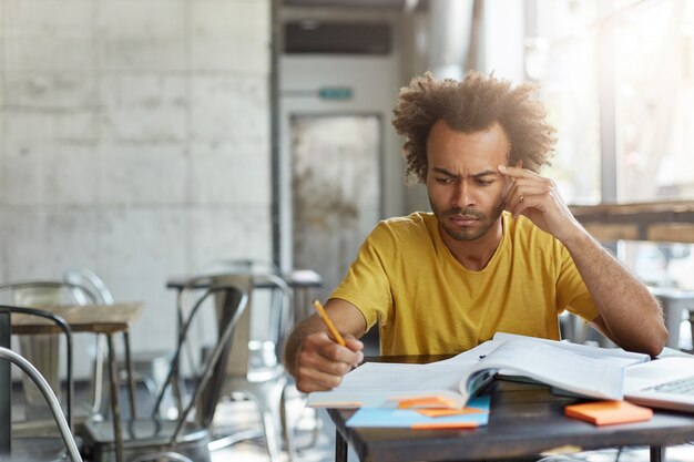 Bello serio studente di carnagione scura che indossa una maglietta gialla che prende appunti con la matita mentre è seduto al tavolo del bar con computer portatile e libri di testo, facendo ricerca