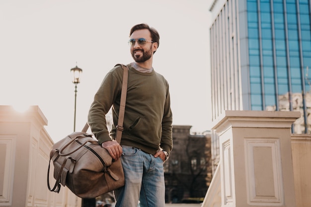 Bello sorridente elegante hipster uomo che cammina in una strada di città con borsa in pelle che indossa felpa e occhiali da sole, tendenza stile urbano, giornata di sole