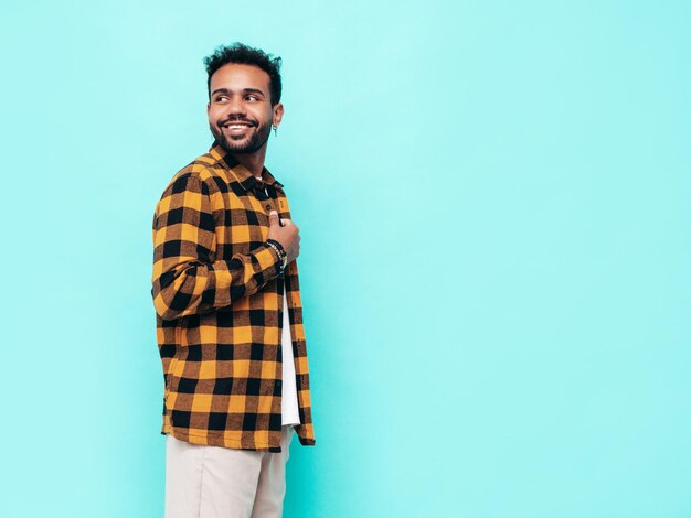 Bello sorridente modello hipster Uomo sexy con la barba lunga vestito in camicia estiva gialla e jeans vestiti Moda maschio in posa vicino al muro blu in studio Allegro e felice Isolato