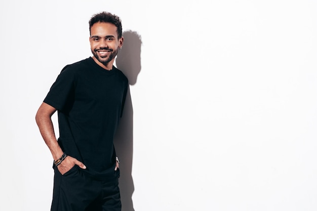 Bello sorridente modello hipster Uomo sexy con la barba lunga vestito in maglietta estiva nera e jeans vestiti Moda maschio in posa vicino al muro bianco in studio al giorno di sole isolato