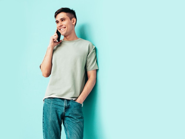 Bello sorridente modelSexy uomo alla moda che parla allo smartphone Moda hipster maschio in posa vicino al muro blu in studio che tiene il telefono con il cellulare isolato