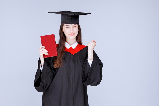 Bello studente che indossa mortarboard che tiene libro rosso. Foto di alta qualità