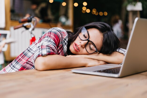 Bello studente dai capelli scuri che dorme vicino al computer. Ritratto all'aperto di affascinante libero professionista femminile in camicia a scacchi restng dopo il lavoro.