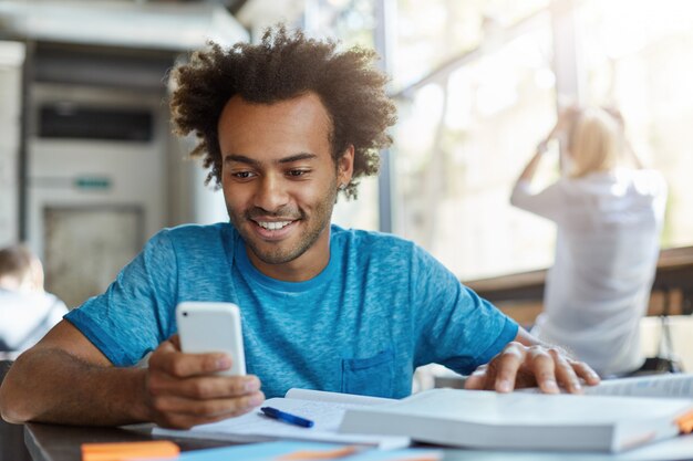 Bello studente gioioso utilizza lo smartphone, guardando lo schermo con un ampio sorriso, seduto al tavolo del bar con un libro di testo e un quaderno