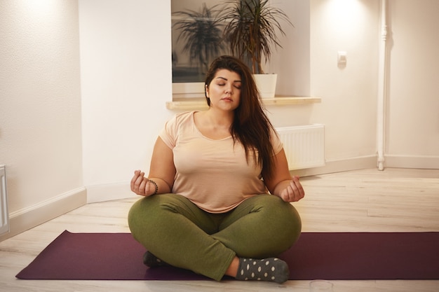 Benessere, armonia, yoga, meditazione, zen e relax. Giovane femmina paffuta obesa seduta sul tappeto, chiudendo gli occhi e tenendo le gambe incrociate, meditando, alla ricerca di pace interiore ed equilibrio