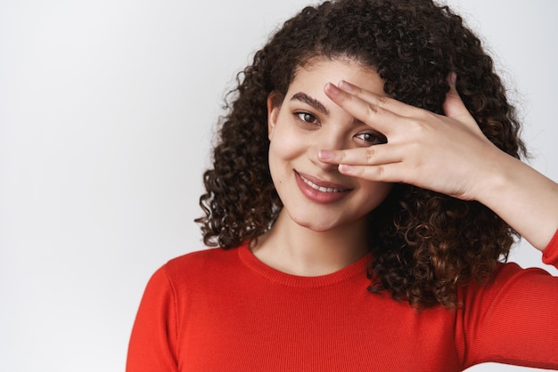 Benessere, felicità e tenerezza concetto Attraente ragazza ispanica acconciatura riccia scura copertura occhio sciocco sguardo attraverso le dita sguardo divertito tenero sorriso gentile in piedi sfondo bianco