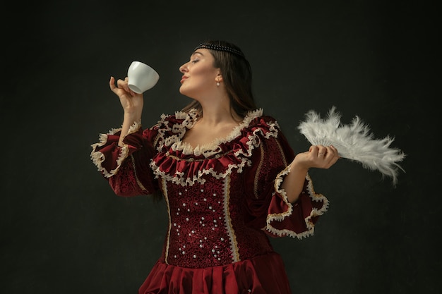 Bere caffè, tiene soffice ventola. Giovane donna medievale in abbigliamento vintage rosso su sfondo scuro. Modello femminile come duchessa, persona reale. Concetto di confronto di epoche, moderno, moda, bellezza.