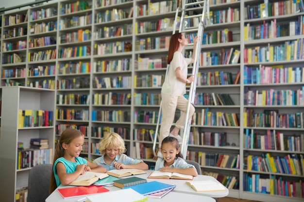 Biblioteca. Bambini allegri della scuola primaria che leggono al tavolo e giovane donna dai capelli lunghi sulle scale vicino allo scaffale per libri in biblioteca