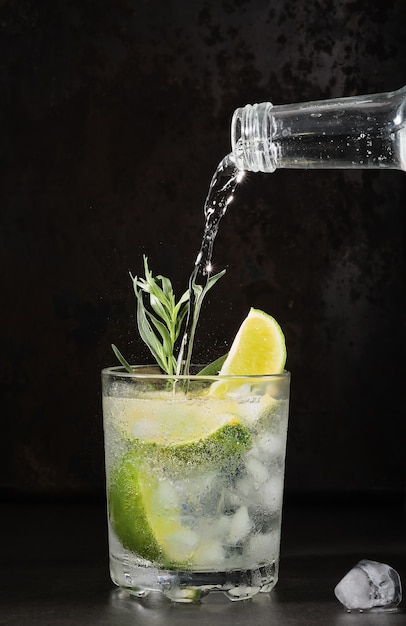 Bicchiere di limonata al lime sul tavolo scuro, bevande estive. L'acqua minerale pura viene versata nel bicchiere. Cornice verticale, messa a fuoco selettiva. Bevanda fatta in casa con lime, dragoncello e cubetti di ghiaccio. Idea di bevande fresche fresche