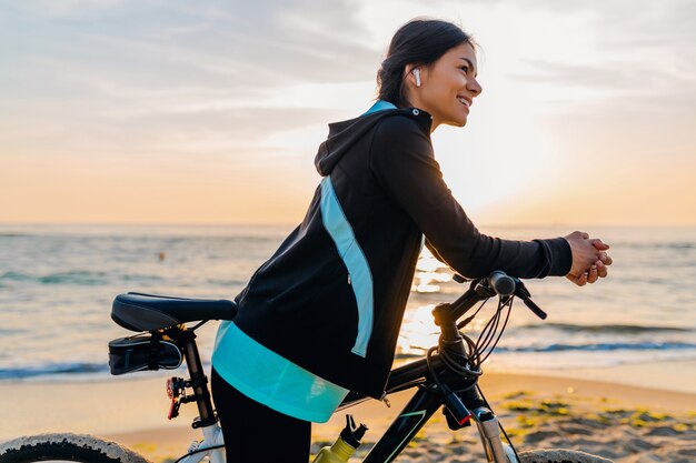 Bicicletta di guida della giovane donna sottile attraente, sport nella spiaggia di estate di alba di mattina nell'usura di idoneità di sport, stile di vita sano attivo, sorridendo felice divertendosi