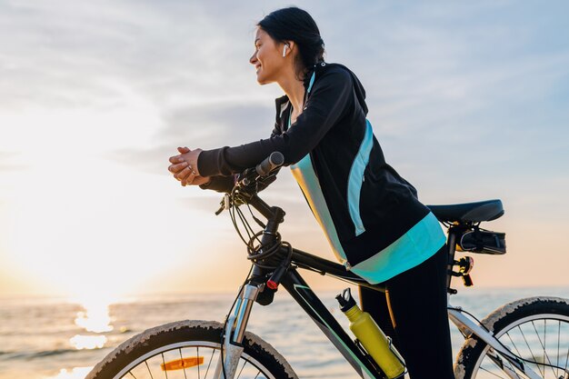 Bicicletta di guida della giovane donna sottile attraente, sport nella spiaggia di estate di alba di mattina nell'usura di idoneità di sport, stile di vita sano attivo, sorridendo felice divertendosi