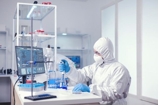 Biologo esperto vestito con pipetta di tenuta in dpi con soluzione blu. Chimico in un moderno laboratorio che fa ricerca utilizzando l'erogatore durante l'epidemia globale con covid-19.