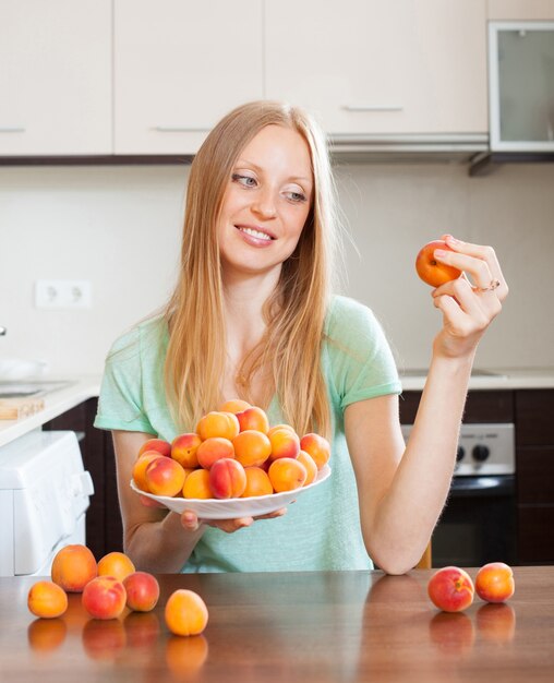 Bionda donna dai capelli lunghi che mangia albicocche nella cucina domestica